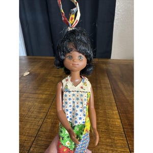 May include: A vintage doll with dark skin, black hair, and brown eyes. The doll wears a patchwork jumpsuit with a white top adorned with blue stars and red accents. A colorful fabric strip is tied in the doll's hair.