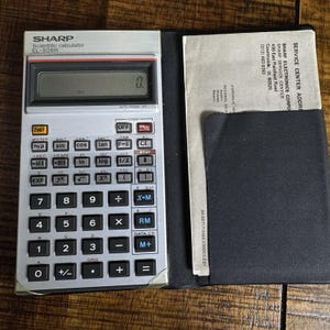 Puede incluir: Calculadora científica Sharp EL-506H plateada con teclas negras y una pantalla digital. La calculadora está abierta, revelando una funda negra y un inserto de papel con texto. La calculadora mide aproximadamente 15 cm de largo.