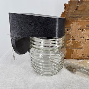 May include: A wall-mounted light fixture featuring a black textured metal housing and a clear, ribbed glass globe. The fixture includes a white electrical wire, a brown cardboard box labeled McGraw-Edison, and a small plastic bag containing metal screws.
