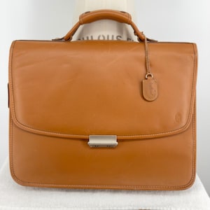 May include: A tan leather briefcase featuring a top handle, a front flap with a silver-toned metal clasp, and a small leather luggage tag attached to the handle. The bag has visible stitching along the edges and a small embossed logo on the front.