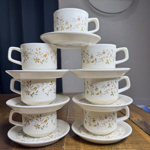 May include: A set of six off-white ceramic cups and saucers stacked in a pyramid formation. Each piece features a speckled finish and a delicate botanical pattern with small yellow, orange, and green flowers and thin brown vines wrapping around.