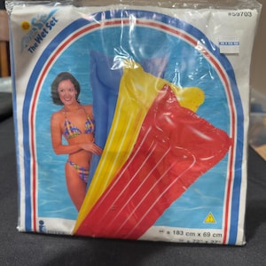 Puede incluir: Un flotador inflable para piscina en azul, amarillo y rojo, empaquetado en plástico transparente. El empaque incluye el texto The Wet Set, #59703 y las dimensiones 183 cm x 69 cm. Un gráfico muestra a una persona en bikini a cuadros.