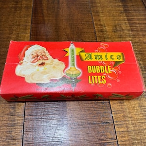 Op de afbeelding: Een rode doos met Amico Bubble Lites kerstverlichting. De doos bevat een illustratie van de kerstman en een bubbellichtafbeelding. De tekst "Amico Bubble Lites" wordt weergegeven in goud en wit.