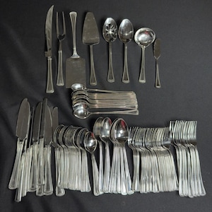 May include: A large collection of silver-toned stainless steel flatware including dinner knives, forks, spoons, a serving fork, a cake server, a slotted spoon, a ladle, and a butter knife. Each piece features a simple, classic handle design.