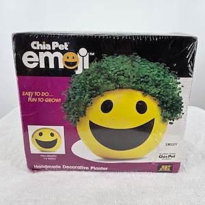 May include: A Chia Pet Emoji Smiley handmade decorative planter in its original packaging. The box features a yellow smiley face with green sprouts growing on top, with text including Chia Pet, emoji, and Easy to do... Fun to grow!.