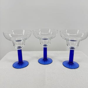 May include: Three clear margarita glasses with a blue stem and base. The glasses have a wide, shallow bowl and a classic margarita shape. The blue base and stem provide a striking contrast to the clear glass.
