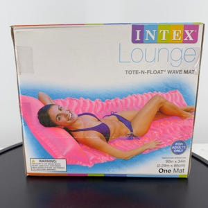 Könnte beinhalten: Ein rechteckiger Karton für eine Intex Lounge Tote-N-Float Wave Matte. Die Verpackung zeigt eine Grafik einer Person, die auf einer leuchtend pinken aufblasbaren Poolmatte liegt. Der Karton enthält den Text Intex Lounge, Tote-N-Float Wave Mat und 2,29m x 86cm.