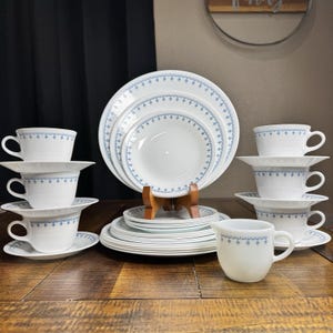 Pode incluir: Um conjunto de louça branco com um padrão geométrico de estrelas azuis nas bordas. A coleção inclui pratos de jantar, pratos de salada, pires e chávenas empilhados, juntamente com uma leiteira, dispostos sobre uma superfície de madeira.