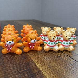 May include: A collection of small, orange teddy bear figurines with red and white polka dot scarves. Some bears wear red, white, and green sweaters. The bears are arranged on a wooden surface.