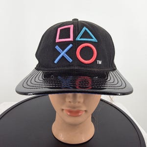 May include: A black baseball cap featuring embroidered PlayStation controller symbols in pink and blue on the front crown. The cap has a structured black crown and a shiny, patent-like black brim with visible stitching details and a small TM symbol.