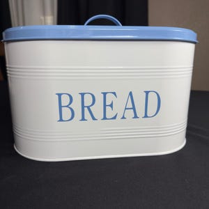 May include: An oval-shaped white metal bread box featuring a light blue lid with a matching arched handle. The word BREAD is printed in large, light blue capital letters on the front of the container, which also has horizontal ribbed detailing.