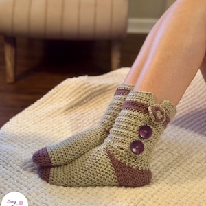 Può includere: Calzini all'uncinetto fatti a mano in beige e malva. I calzini presentano un design a coste, dettagli malva e bottoni viola decorativi. Il polsino è adornato da un piccolo fiore. Progettati per il comfort e il calore.