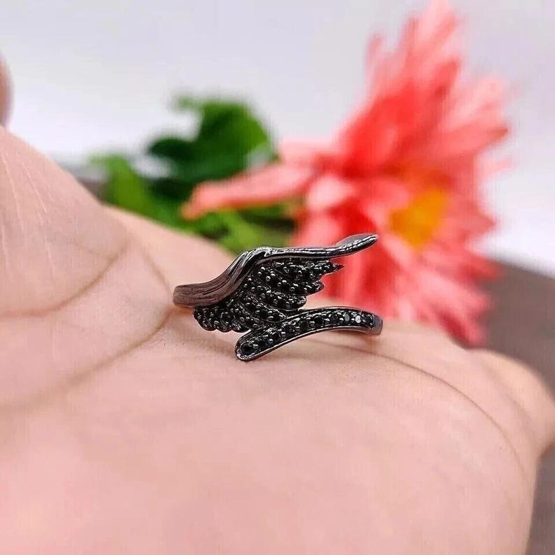 May include: A black ring with a wing design, adorned with small black stones. The ring's wing detail curves upwards, creating a unique and stylish accessory. The ring is a dark color, and the stones add a touch of sparkle.