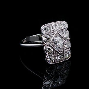 May include: A silver-toned ring featuring a rectangular head set with multiple round-cut clear stones arranged in a geometric pattern, centered around a larger oval-shaped stone, with a smooth, thin band and a small dark rectangular mark on the side.