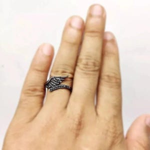 May include: A black ring with a wing design. The ring features a wing shape with small, dark stones. The ring is worn on a finger, showcasing the intricate details and dark color of the jewelry.