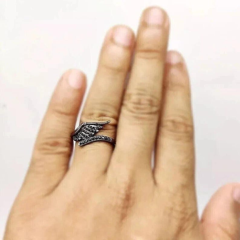 May include: A black ring with a wing design. The ring features a wing shape with small, dark stones. The ring is worn on a finger, showcasing the intricate details and dark color of the jewelry.