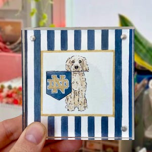 Peut inclure: Un bloc acrylique carr&eacute; avec une illustration &agrave; l'aquarelle d'un chien duveteux et un bouclier bleu et or avec les lettres "ND". Le bloc a une bordure ray&eacute;e bleue et blanche. Le bloc est tenu par une main.