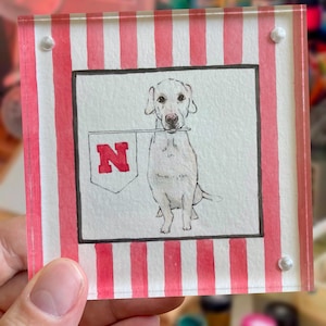 Peut inclure: Un bloc carr&eacute; en acrylique avec une peinture &agrave; l'aquarelle d'un chien blanc tenant une banni&egrave;re avec un "N" rouge. Le fond pr&eacute;sente des rayures verticales rouges et blanches. Le bloc est tenu par une main.
