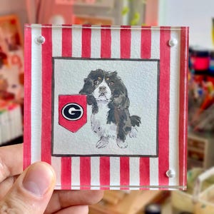 Peut inclure: Une peinture &agrave; l'aquarelle d'un chien Cavalier King Charles Spaniel tenant un fanion rouge et noir avec un fond blanc et la lettre "G". La peinture est plac&eacute;e sur un fond &agrave; rayures rouges et blanches, encadr&eacute;e dans un cadre en acrylique transparent.