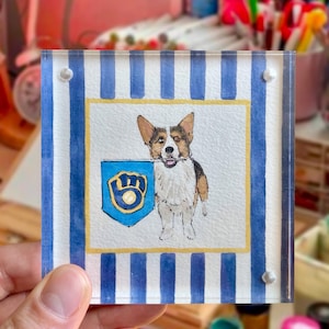 Peut inclure: Sous-verre carr&eacute; en acrylique avec une peinture &agrave; l'aquarelle d'un chien Corgi tenant un bouclier bleu avec un embl&egrave;me dor&eacute;. Le dessous de verre a des rayures verticales bleues et blanches et est maintenu par quatre rivets argent&eacute;s.