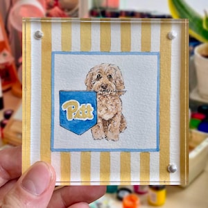 Peut inclure: Un bloc acrylique carr&eacute; avec une peinture &agrave; l'aquarelle d'un chien brun clair et duveteux tenant un fanion bleu avec le mot "Pitt". Le fond pr&eacute;sente des rayures verticales jaunes et blanches, et la peinture est fix&eacute;e avec des rivets argent&eacute;s.