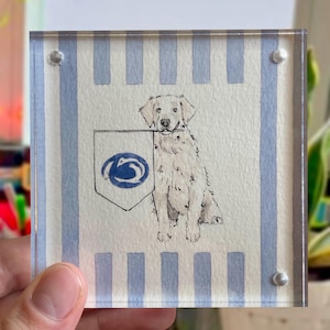Peut inclure: Un bloc acrylique carr&eacute; avec une illustration &agrave; l'aquarelle d'un golden retriever tenant un fanion avec un logo bleu. Le fond pr&eacute;sente des rayures verticales bleues et blanches. L'&oelig;uvre est maintenue par quatre rivets argent&eacute;s.