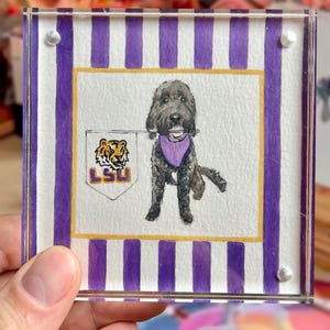 Peut inclure: Un bloc acrylique carr&eacute; avec une peinture &agrave; l'aquarelle d'un chien noir portant un bandana violet. Le chien tient une banni&egrave;re LSU. Le fond pr&eacute;sente des rayures violettes et blanches. Le bloc est tenu par une main.