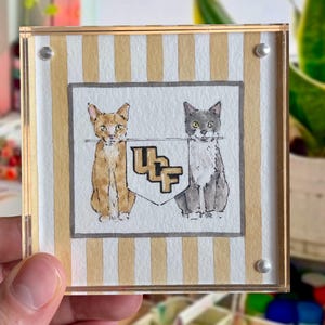 Peut inclure: Un dessous de verre carr&eacute; en acrylique avec une peinture &agrave; l'aquarelle de deux chats encadrant un &eacute;cusson avec les lettres "UF". Le fond pr&eacute;sente des rayures verticales dor&eacute;es et blanches. Le dessous de verre est tenu par une main.
