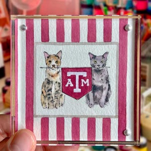 Peut inclure: Sous-verre carr&eacute; en acrylique avec une peinture &agrave; l'aquarelle de deux chats encadrant une banni&egrave;re bordeaux et blanche avec les lettres "A M". Le fond pr&eacute;sente des rayures roses et blanches. Le dessous de verre est tenu par une main.