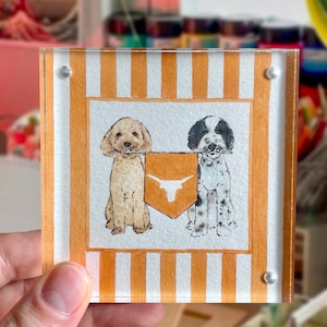 Puede incluir: Posavasos cuadrado de acrílico con ilustraciones de dos perros sosteniendo un escudo con un logotipo de Longhorn. El posavasos tiene un borde a rayas naranja y blanco y está sujeto con remaches plateados.