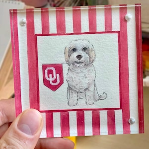 Peut inclure: Peinture &agrave; l'aquarelle d'un chien blanc tenant un drapeau rouge avec un logo "Q" blanc. La peinture est plac&eacute;e sur un fond de rayures rouges et blanches, encadr&eacute;e dans un cadre acrylique transparent avec des vis argent&eacute;es.