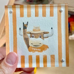 Könnte beinhalten: Quadratischer Acryl-Untersetzer mit einer Aquarellillustration eines Longhorn-Stiers, der einen Hut und ein Hemd mit dem Wort "TEXAS" trägt. Der Untersetzer hat orange und weiße Streifen und wird von einer Hand gehalten.