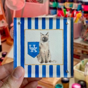 Peut inclure: Un bloc carr&eacute; en acrylique avec une peinture &agrave; l'aquarelle d'un chat siamois tenant une banni&egrave;re bleue avec un logo "UK" blanc. Le bloc a des bordures &agrave; rayures bleues et blanches et est tenu par une main.