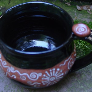 May include: A black ceramic mug with a brown rim and white swirl designs. The mug has a handle with a brown knob.