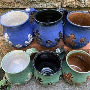 May include: Six handmade ceramic mugs in shades of blue and green. The mugs feature floral and poodle designs, with a variety of interior colors. Each mug has a unique handle design with decorative accents. The mugs are arranged on a stone and brick surface.