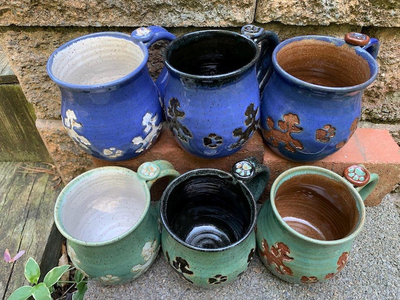 May include: Six handmade ceramic mugs in shades of blue and green. The mugs feature floral and poodle designs, with a variety of interior colors. Each mug has a unique handle design with decorative accents. The mugs are arranged on a stone and brick surface.
