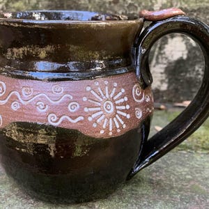 May include: A brown ceramic mug with a white design of swirls and a sunburst. The mug has a rounded body and a handle.