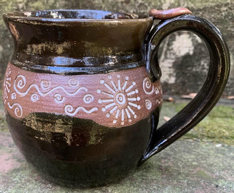 May include: A brown ceramic mug with a white design of swirls and a sunburst. The mug has a rounded body and a handle.
