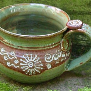 May include: A green and brown ceramic mug with a unique handle and white swirl designs. The mug has a rustic, handmade look.