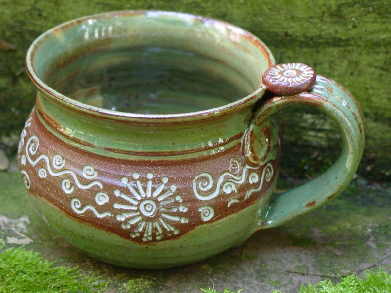 May include: A green and brown ceramic mug with a unique handle and white swirl designs. The mug has a rustic, handmade look.