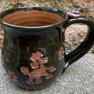 May include: A handmade ceramic mug with a dark green exterior and a brown interior. The mug features embossed brown poodle designs around the body and a matching accent on the handle. The mug has a glossy finish.