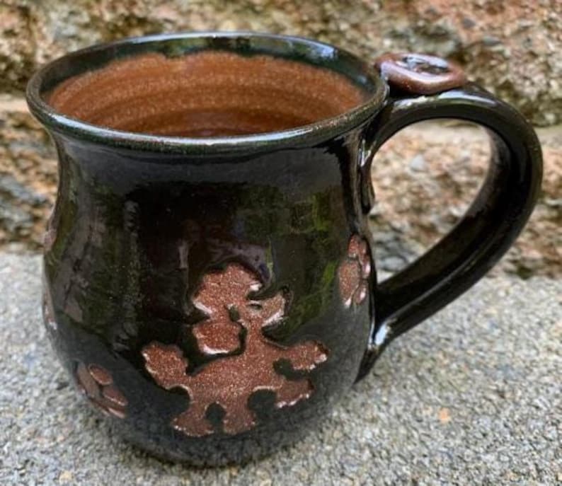 May include: A handmade ceramic mug with a dark green exterior and a brown interior. The mug features embossed brown poodle designs around the body and a matching accent on the handle. The mug has a glossy finish.