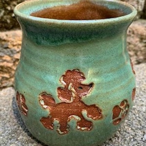 May include: Handmade ceramic vase with a turquoise glaze and brown poodle designs. The interior is a warm brown color. The vase has a rounded shape and a small handle. A unique piece for home decor.
