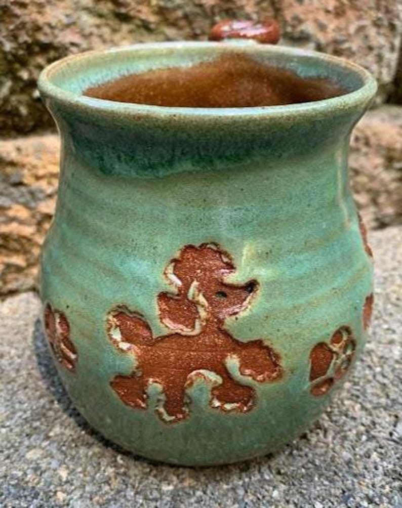 May include: Handmade ceramic vase with a turquoise glaze and brown poodle designs. The interior is a warm brown color. The vase has a rounded shape and a small handle. A unique piece for home decor.