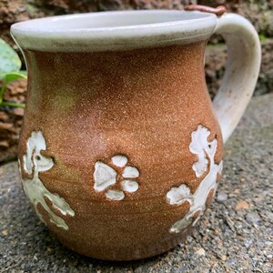 May include: Brown ceramic mug with a white rim and handle. The mug features embossed white poodle designs and a paw print. The mug is handmade and has a rustic, textured appearance, perfect for coffee or tea.