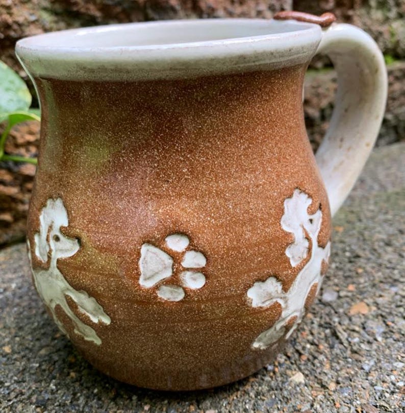 May include: Brown ceramic mug with a white rim and handle. The mug features embossed white poodle designs and a paw print. The mug is handmade and has a rustic, textured appearance, perfect for coffee or tea.