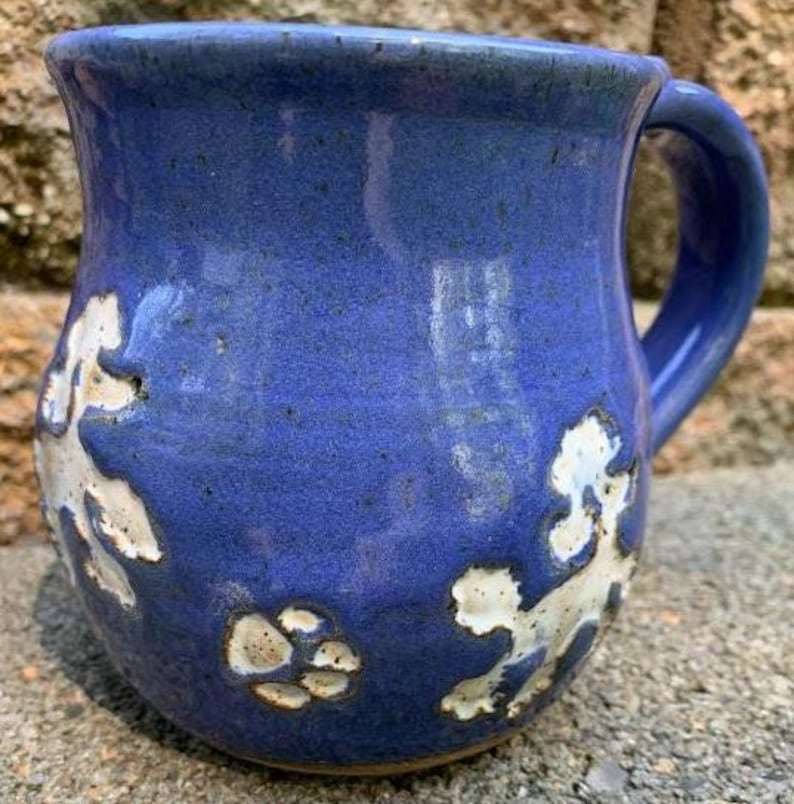 May include: A handmade ceramic mug with a deep blue glaze and a white floral design. The mug has a rounded shape with a handle and is suitable for hot or cold beverages. The mug is approximately 4 inches tall.