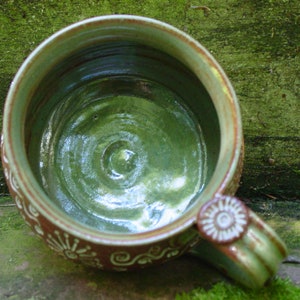 May include: A green ceramic mug with a brown rim and white decorative designs. The inside of the mug is glazed in a green color.