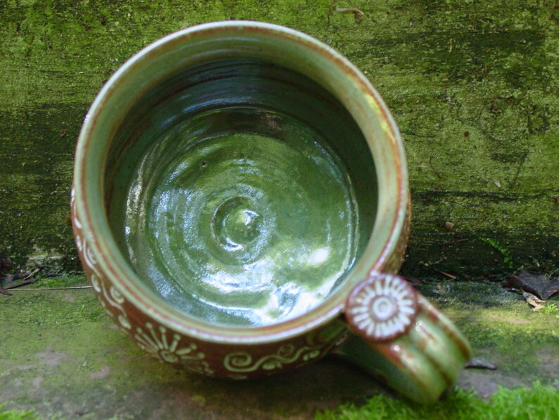 May include: A green ceramic mug with a brown rim and white decorative designs. The inside of the mug is glazed in a green color.