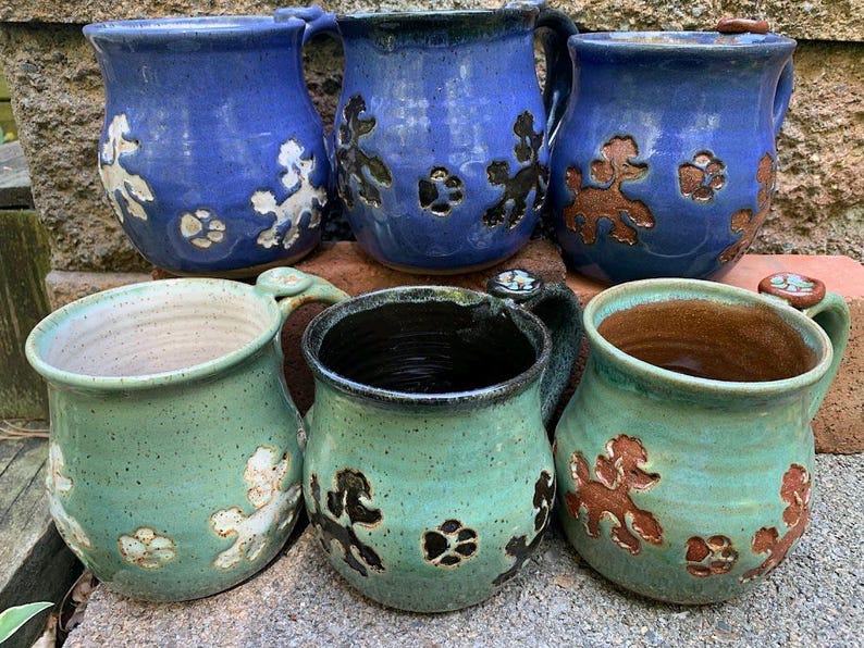 May include: Six handmade ceramic mugs in shades of blue and green. Each mug features a poodle design and paw prints. The mugs have handles and are displayed on a stone surface.